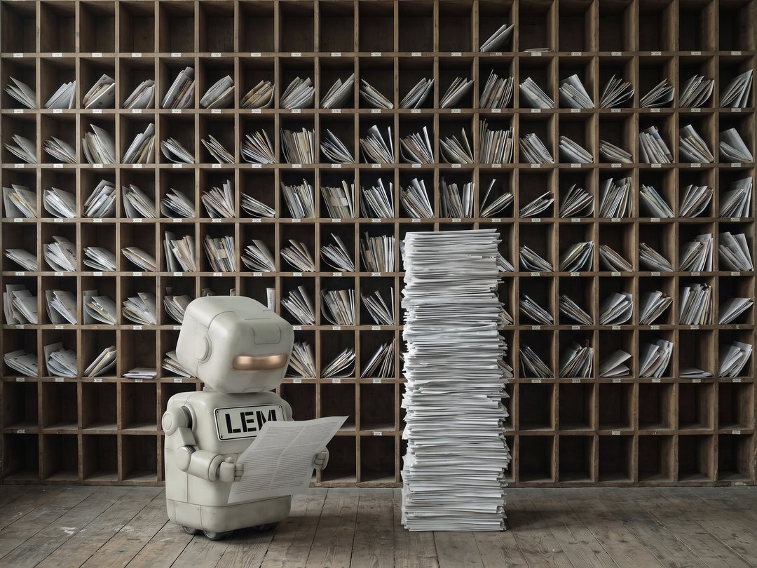 Lem, a small boxy robot with a large LEM name plate across their front, reads a single opened letter in front of a floor-to-ceiling wall of pigeonholes stuffed with post, beside a stack of unopened letters taller than they are.